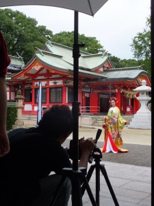 「大分のウェディングBOOK　ハピマリ」の衣裳撮影の風景。艶やかな色打掛が赤い社殿をバックにしてより映えます。